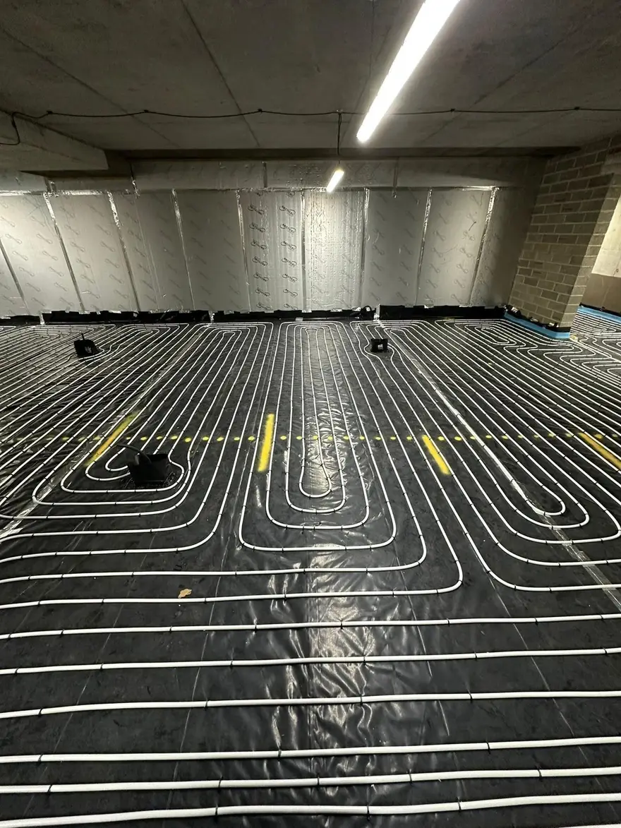 A large room with a concrete ceiling and foil faced wall insulation, its floor covered edge to edge with white underfloor heating pipe clipped in even loops over black insulation, ready for screeding.