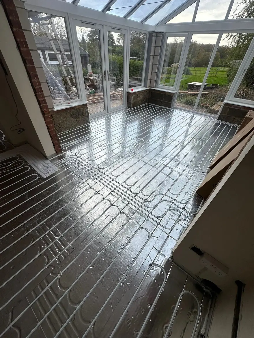 A white uPVC garden room with a glazed roof looking onto a garden, its floor covered with silver foil faced grooved panels and underfloor heating pipe pressed into the channels.