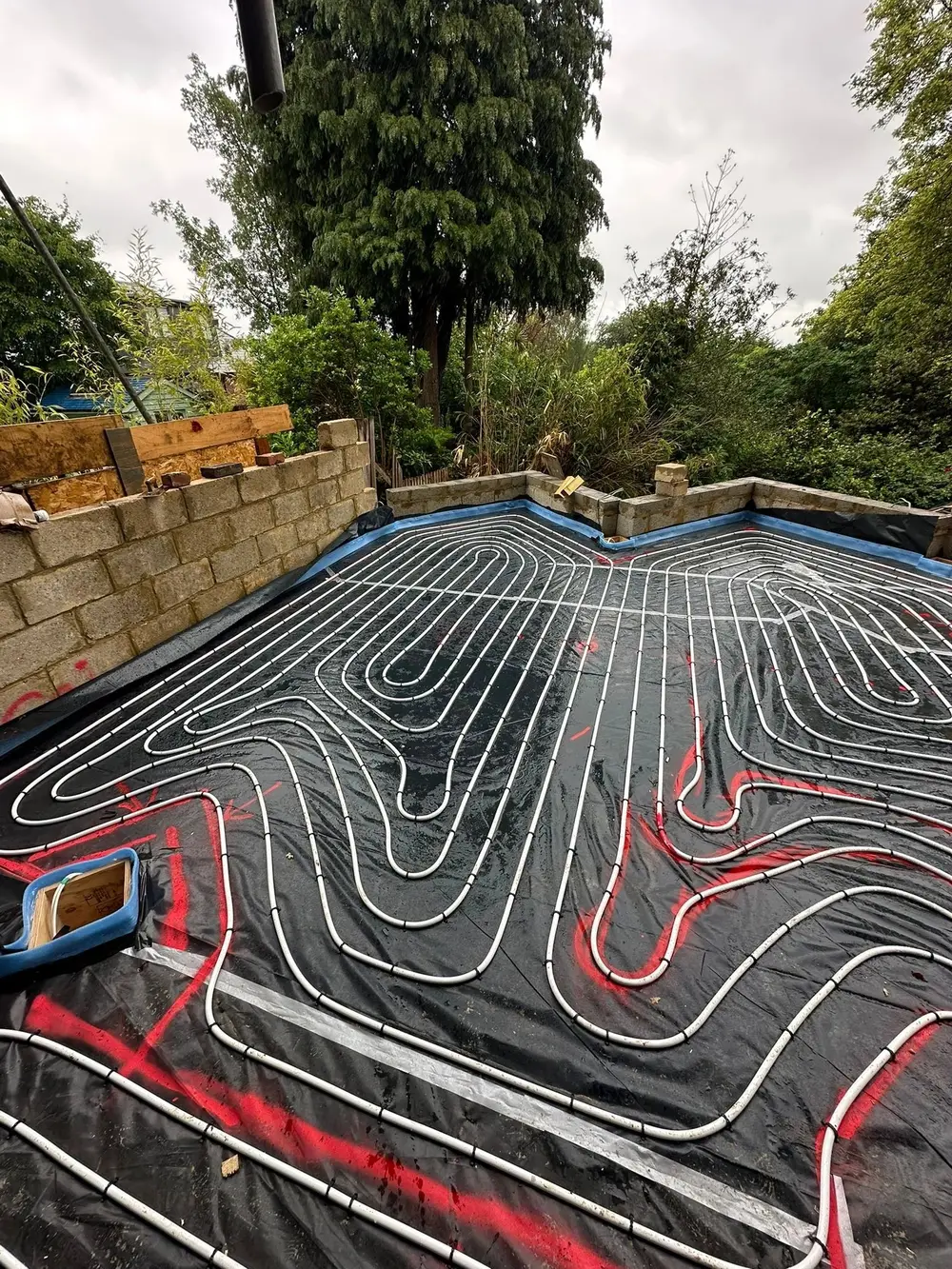 A ground floor extension under construction with blockwork walls, its floor covered with membrane and underfloor heating pipe laid in sweeping loops with layout marks sprayed on.