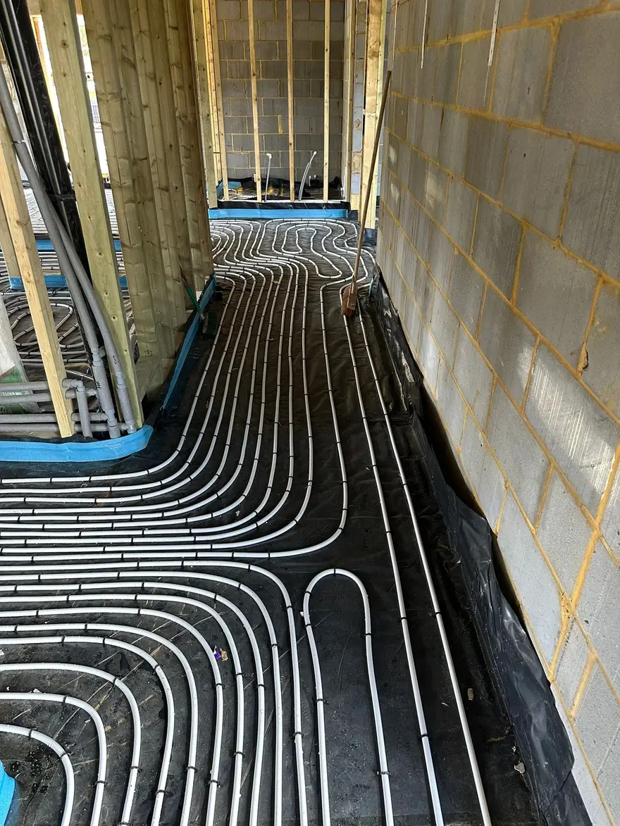A building project at first fix stage with timber stud walls and blockwork, its floor covered in black membrane with underfloor heating pipe laid in long curving loops running through several rooms.