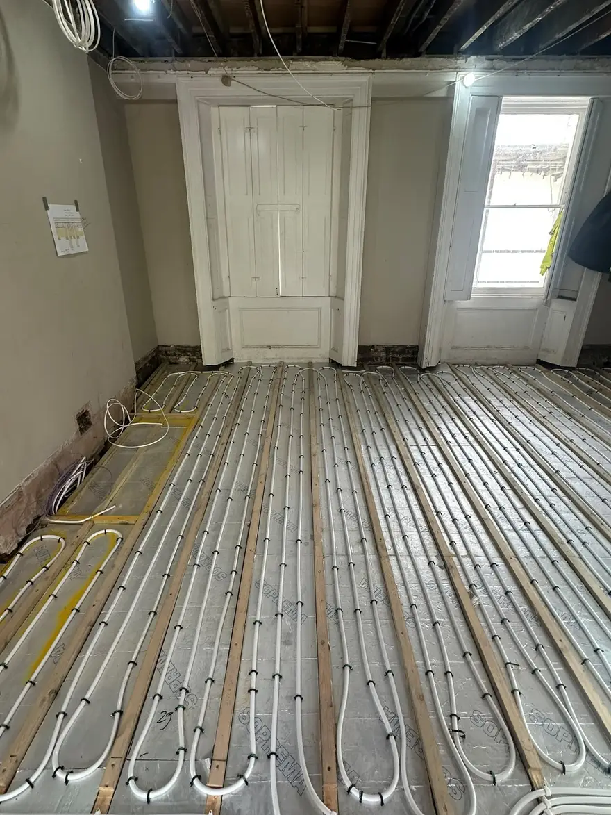 White underfloor heating pipe laid in long parallel loops between timber floor joists over foil faced insulation, across a room with tall shuttered windows and exposed ceiling joists above.